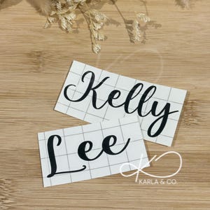 May include: Two white rectangular stickers with black cursive text reading "Kelly" and "Lee". The stickers are on a grid pattern. The background is a wooden surface with dried flowers.