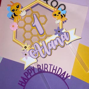 K&ouml;nnte beinhalten: Ein lila und gelber Tortendekor mit einem Wabenmuster, zwei Cartoon-Bienen, einer Eins und dem Namen "Alana" mit dem Text "Happy Birthday".