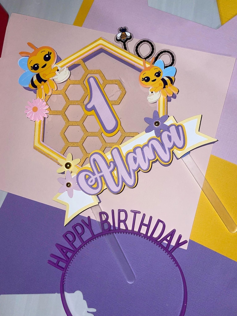 K&ouml;nnte beinhalten: Ein lila und gelber Tortendekor mit einem Wabenmuster, zwei Cartoon-Bienen, einer Eins und dem Namen "Alana" mit dem Text "Happy Birthday".