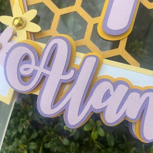 K&ouml;nnte beinhalten: Ein mehrschichtiger Papierschnitt des Namens "Alan" in Lila und Gelb mit einem Wabenmuster im Hintergrund.