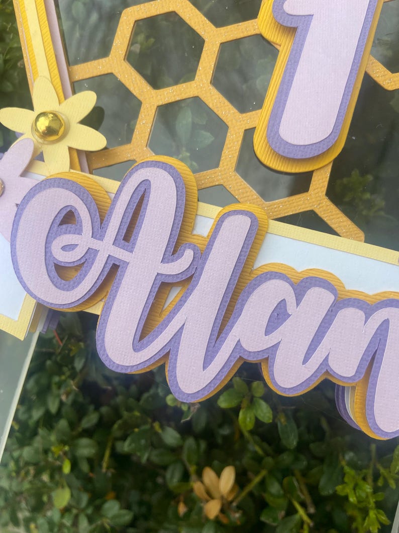 K&ouml;nnte beinhalten: Ein mehrschichtiger Papierschnitt des Namens "Alan" in Lila und Gelb mit einem Wabenmuster im Hintergrund.