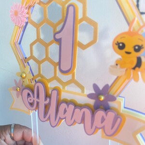 K&ouml;nnte beinhalten: Ein rosa-gelber Kuchentopper im Bienenwaben-Design mit der Zahl "1" und dem Namen "Alana" in rosa Schrift. Der Topper zeigt eine Biene und ein Bienenwabenmuster.