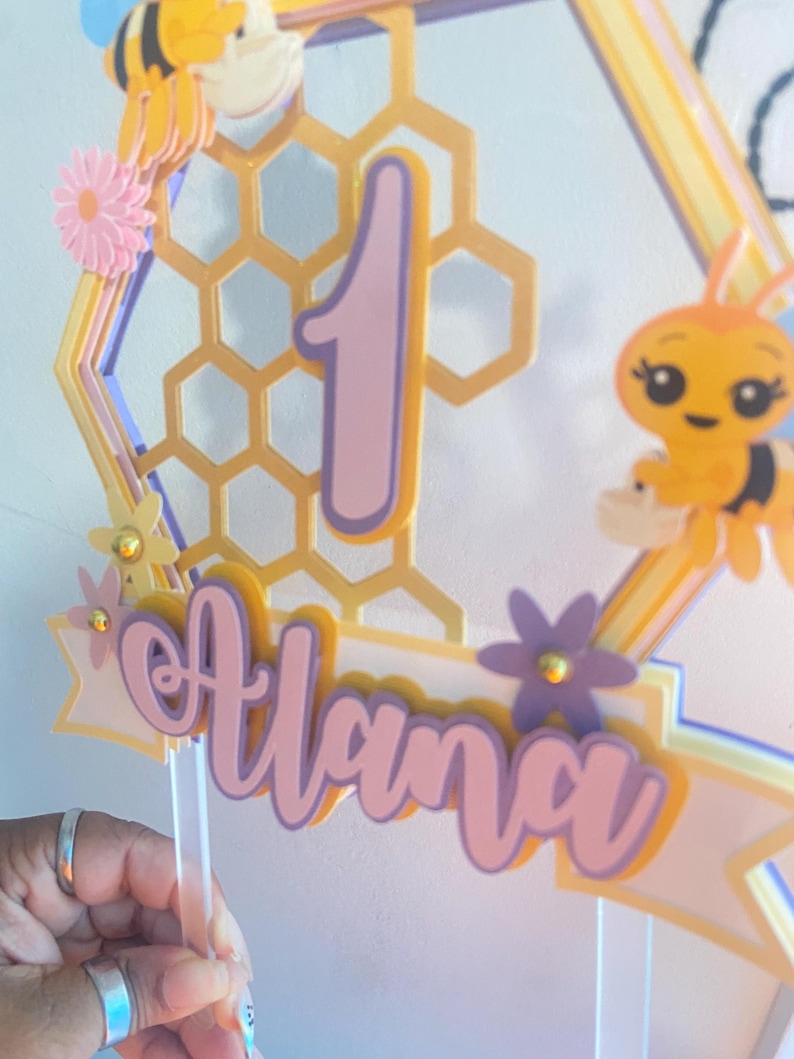 K&ouml;nnte beinhalten: Ein rosa-gelber Kuchentopper im Bienenwaben-Design mit der Zahl "1" und dem Namen "Alana" in rosa Schrift. Der Topper zeigt eine Biene und ein Bienenwabenmuster.