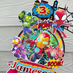 May include: A colorful cake topper featuring superhero characters, including Iron Man, Spider-Man, and the Hulk. The topper has the name "Jameson" and the number "3" displayed. The design includes comic book style "POW!" and "BOOM!" text.