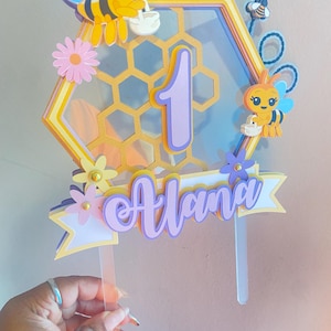 K&ouml;nnte beinhalten: Ein gelber und violetter Waben-Kuchentopper mit einer Biene und der Zahl eins. Der Topper enth&auml;lt auch den Namen "Alana" in violetten Buchstaben.