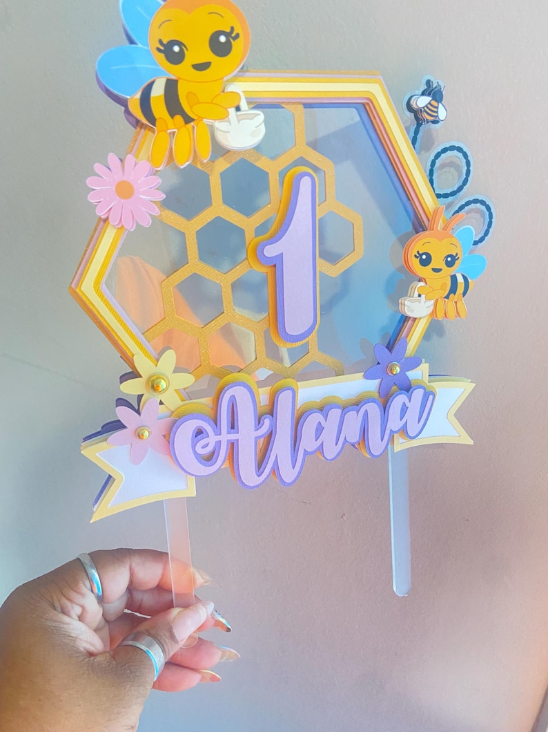 K&ouml;nnte beinhalten: Ein gelber und violetter Waben-Kuchentopper mit einer Biene und der Zahl eins. Der Topper enth&auml;lt auch den Namen "Alana" in violetten Buchstaben.