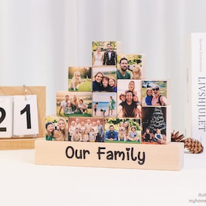 Peut inclure: Un présentoir en blocs de bois avec des photos de famille disposées en gradins. La base porte l'inscription "Our Family" en écriture noire. Un calendrier en bois affiche "JUL 21". Des livres et une pomme de pin sont en arrière-plan.