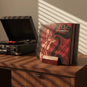 May include: A black portable record player with a red vinyl record is next to a stack of vinyl records. The album cover features a woman with long red hair. A small red sign reads "NOW PLAYING". The records are on a wooden surface.
