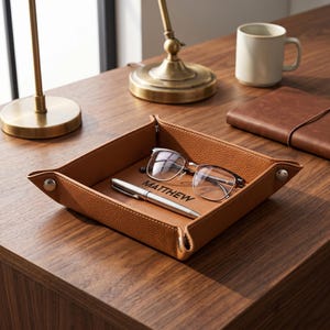 Pode incluir: Uma bandeja de couro castanho com uma caneta prateada, óculos e o nome "MATTHEW" gravado. A bandeja está sobre uma superfície de madeira, com uma caneca e um caderno no fundo.