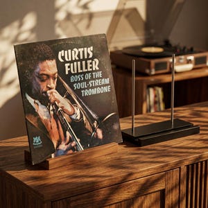 Puede incluir: Una portada de álbum de vinilo vintage de "Boss of the Soul-Stream Trombone" de Curtis Fuller. El álbum se muestra en un soporte de madera. Un soporte de discos negro y un tocadiscos están en el fondo.