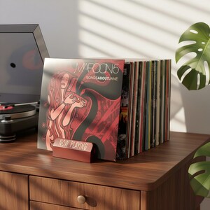 Op de afbeelding: Een stapel vinylplaten met het Maroon 5 album "Songs About Jane" prominent aanwezig. Een rode "Now Playing" standaard toont het album. De platen staan op een houten kast naast een platenspeler.