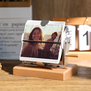 Peut inclure: Un calendrier photo à bascule en bois avec un cadre en métal et une base en bois. Le calendrier affiche une photo d'un couple souriant sur un bateau. Le calendrier est sur une surface en bois.