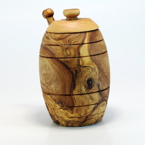 Op de afbeelding: Een houten honingpot in de vorm van een vat met een deksel en een houten roerstaaf. De honingpot is gemaakt van olijfhout en heeft een natuurlijke houtkleur.