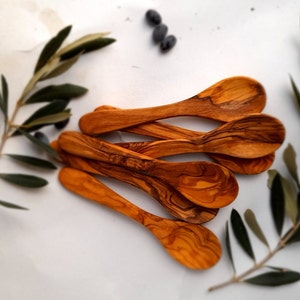 Può includere: Un set di sei cucchiai in legno di ulivo, ognuno con un motivo di venatura unico. I cucchiai sono disposti a ventaglio su uno sfondo bianco.