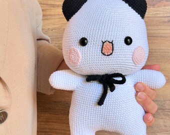 Può includere: Un peluche a forma di panda realizzato a mano all'uncinetto. Il panda è principalmente bianco con dettagli neri sulle orecchie, sui piedi e un fiocco. Ha guance rosa e una piccola bocca ricamata. L'orso è tenuto da una persona.