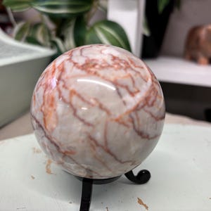 Puede incluir: Una esfera de mármol blanco y rojo con un patrón de remolino, sentada sobre un soporte de metal negro.