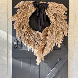 May include: A dried pampas grass wreath with a black satin bow, hanging on a dark grey wooden door.
