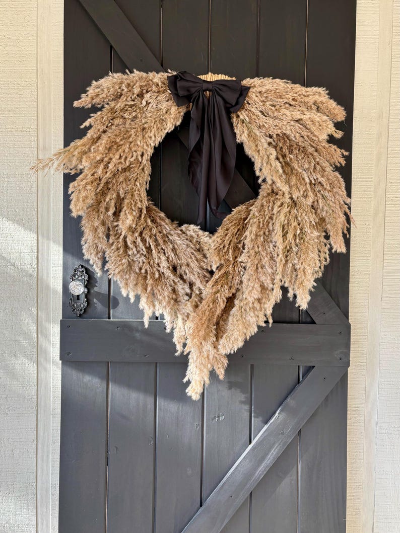 May include: A dried pampas grass wreath with a black satin bow, hanging on a dark grey wooden door.