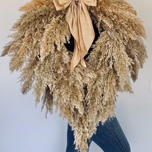 May include: A heart-shaped wreath made of dried pampas grass with a large, beige satin bow tied in the centre.