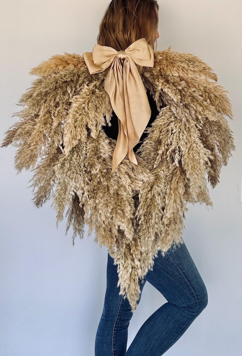 May include: A heart-shaped wreath made of dried pampas grass with a large, beige satin bow tied in the centre.