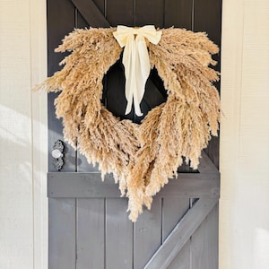 May include: A dried pampas grass wreath with a cream-coloured bow hanging on a dark grey wooden door.