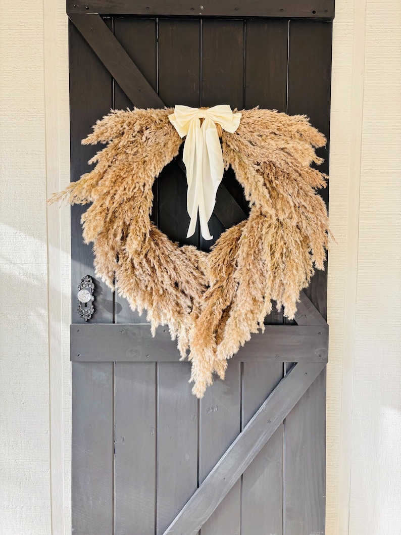 May include: A dried pampas grass wreath with a cream-coloured bow hanging on a dark grey wooden door.