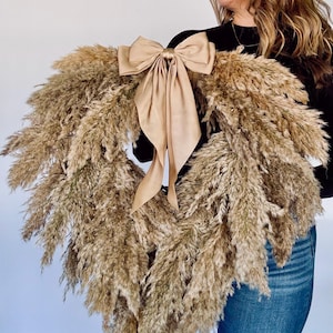 May include: A large pampas grass wreath with a large, beige satin bow. The wreath is being held by a person wearing a black top and blue jeans.