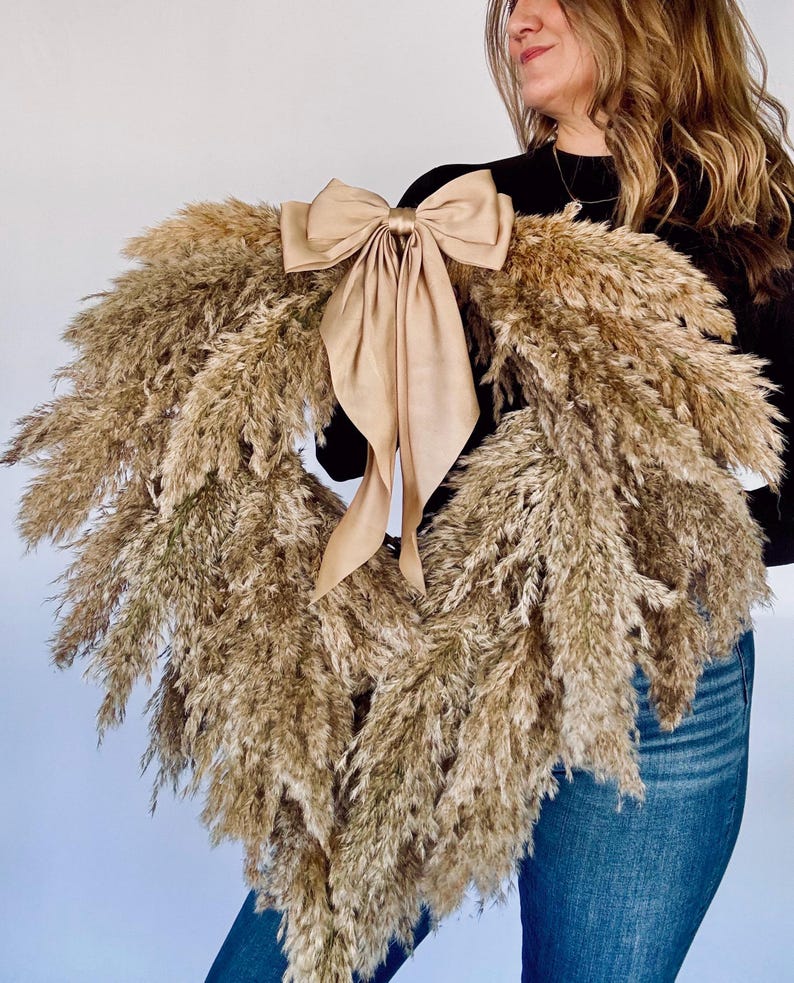 May include: A large pampas grass wreath with a large, beige satin bow. The wreath is being held by a person wearing a black top and blue jeans.