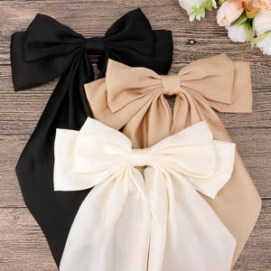 May include: Three large satin bows in black, beige, and white. The bows are tied with long tails and are arranged in a stack on a wooden surface.