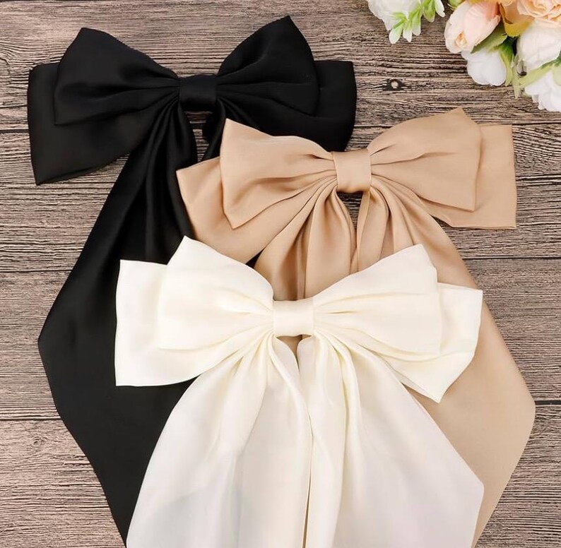 May include: Three large satin bows in black, beige, and white. The bows are tied with long tails and are arranged in a stack on a wooden surface.