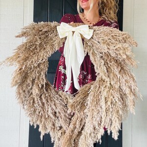 May include: A large pampas grass wreath with a white satin bow. The wreath is being held by a person wearing a burgundy floral dress.
