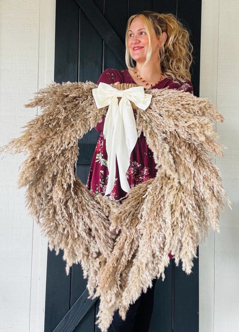 May include: A large pampas grass wreath with a white satin bow. The wreath is being held by a person wearing a burgundy floral dress.