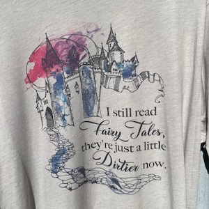 Op de afbeelding: Een lichtgrijs T-shirt met een aquarel-illustratie van een kasteel en de tekst "I still read Fairy Tales, they're just a little Dirtier now."