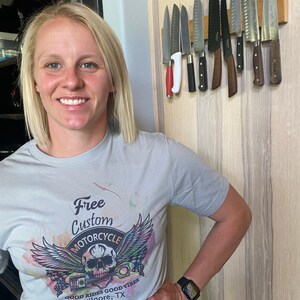 May include: A woman wearing a grey t-shirt with a colourful graphic of a skull with wings and the text "Free Custom Motorcycle, Good Rides Good Vibes, Kilgore, TX, L.V."
