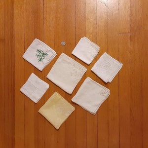 May include: Eight white handkerchiefs with embroidered designs, one with a green floral design, on a wooden surface.