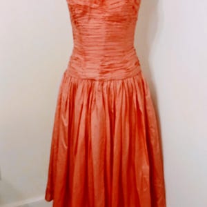 May include: A coral-colored dress on a mannequin. The bodice is ruched, and the skirt is gathered, creating a full silhouette. The dress has thin spaghetti straps and a sweetheart neckline. The dress is displayed on a vintage dress form.