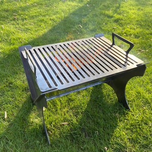May include: A black metal grill with a rectangular cooking surface and two legs. The grill is sitting on a grassy lawn.