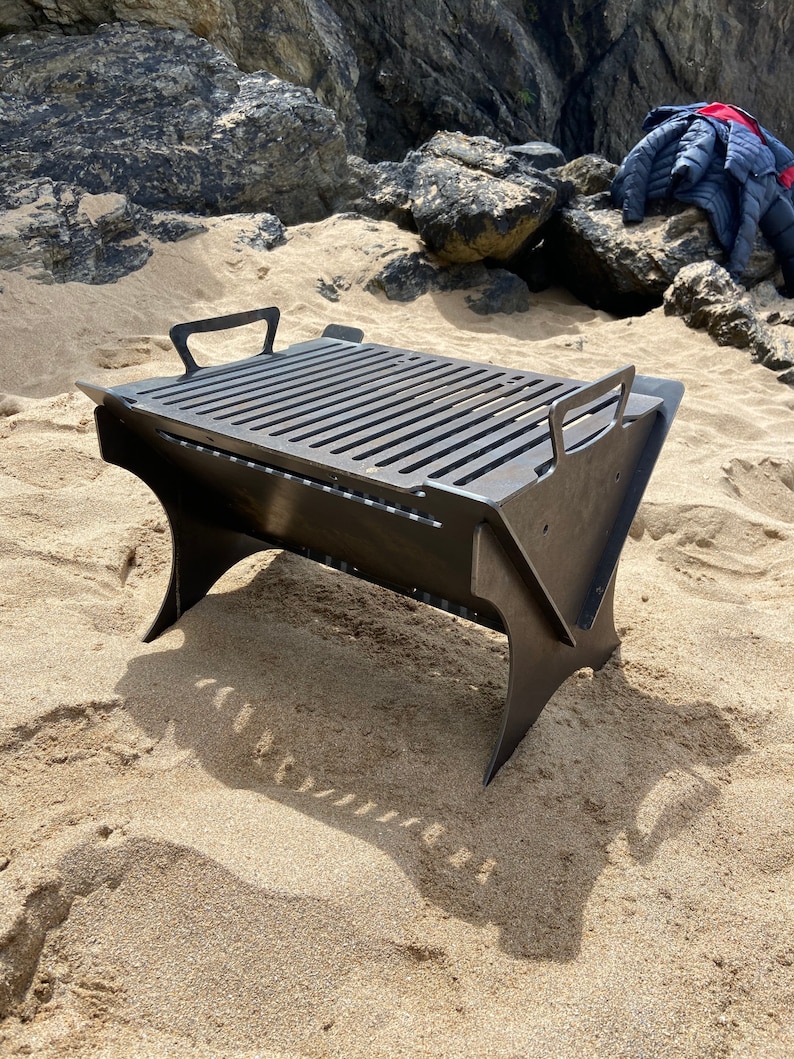 May include: A black metal folding grill with a rectangular cooking surface and a handle on each side. The grill is sitting on a sandy beach.