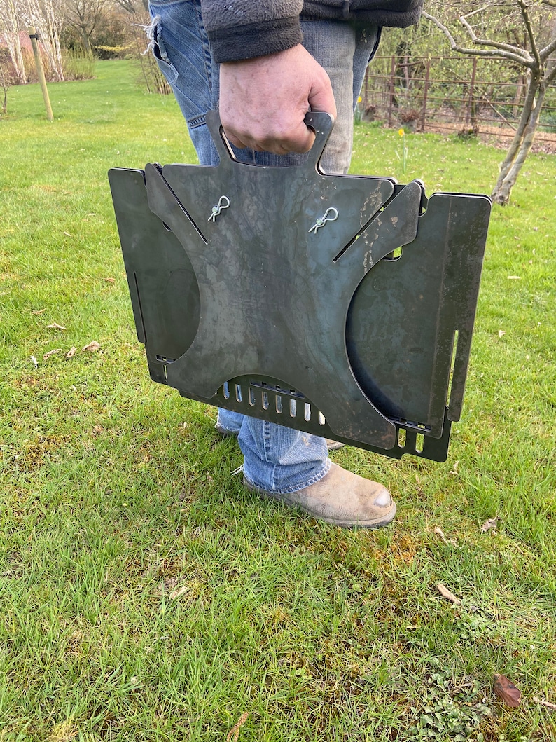 May include: A portable, foldable metal fire pit with a handle. The fire pit is made of dark metal and has a rustic look. It is folded up and being held by a person.