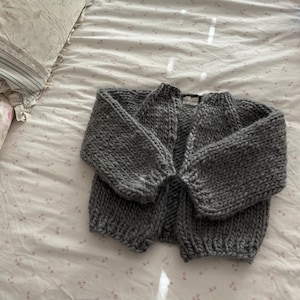 May include: A gray knitted cardigan with a simple design. The cardigan is open in the front and has long sleeves.
