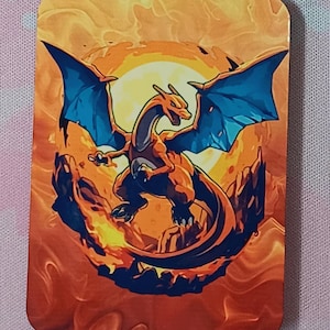 May include: Rectangular orange and yellow graphic featuring a Charizard, a dragon-like Pokémon, with blue wings, set against a fiery background. The artwork depicts the Pokémon in a dynamic pose, with a bright sun in the background.