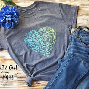 May include: A gray t-shirt with a heart-shaped design featuring the words "Neurofibromatosis Type II" and other related terms. The shirt is displayed on a wooden surface with a pair of blue jeans.
