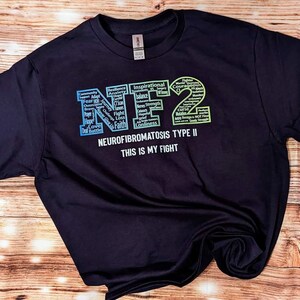 Può includere: Maglietta nera con un design a gradiente arcobaleno che scrive "NF2" con parole come "speranza", "fede", "amore" e "forza" all'interno delle lettere. Il testo "Neurofibromatosi di tipo II Questa è la mia battaglia" è stampato sotto il design.