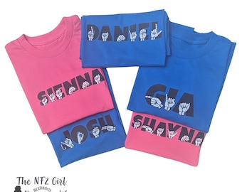 May include: Five t-shirts with names spelled out in American Sign Language. The names are Daniel, Sienna, Gia, Josh, and Shayna. The shirts are pink and blue.