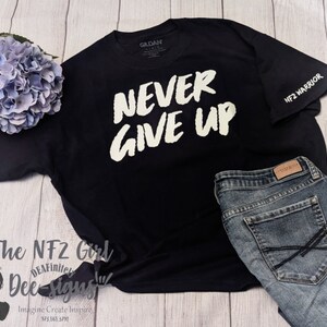 Può includere: Maglietta nera con testo bianco che recita "NEVER GIVE UP". La maglietta ha una piccola toppa NF2 WARRIOR sulla manica.