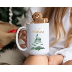 Op de afbeelding: Een witte keramische mok met een groen kerstboomontwerp en de tekst "Merry Christmas". Een gingerbread man koekje zit bovenop de mok.