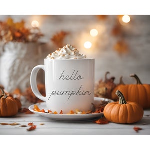 May include: White ceramic mug with the text "hello pumpkin" on it. The mug is filled with a creamy beverage topped with whipped cream and cinnamon. The mug is sitting on a white surface with autumn leaves and pumpkins.