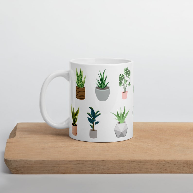 May include: White ceramic mug featuring a repeating pattern of potted plants in various shapes and sizes. The plants are depicted in green, brown, and gray, with a minimalist design. The mug is placed on a wooden surface.
