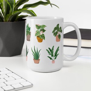 May include: White ceramic mug featuring illustrations of various potted plants in shades of green, pink, and brown. The plants include succulents, ferns, and other foliage. The mug has a large handle and is suitable for hot or cold beverages.