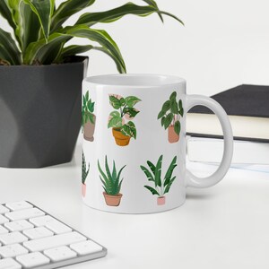 May include: White ceramic mug featuring illustrations of various potted plants in shades of green, pink, and brown. The mug has a curved handle and is set against a neutral background with a keyboard and greenery.